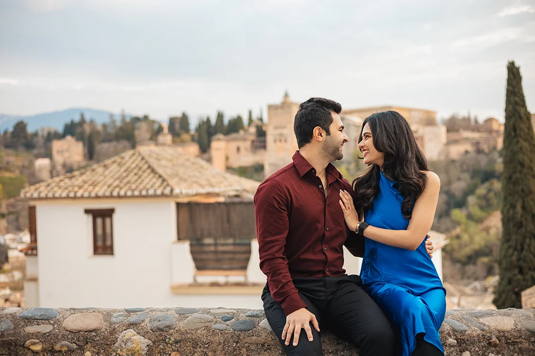 A romantic couple shares a smile while sitting on a stone ledge overlooking the historic Alhambra palace in Granada, showcasing professional photography in the Albaicín district.