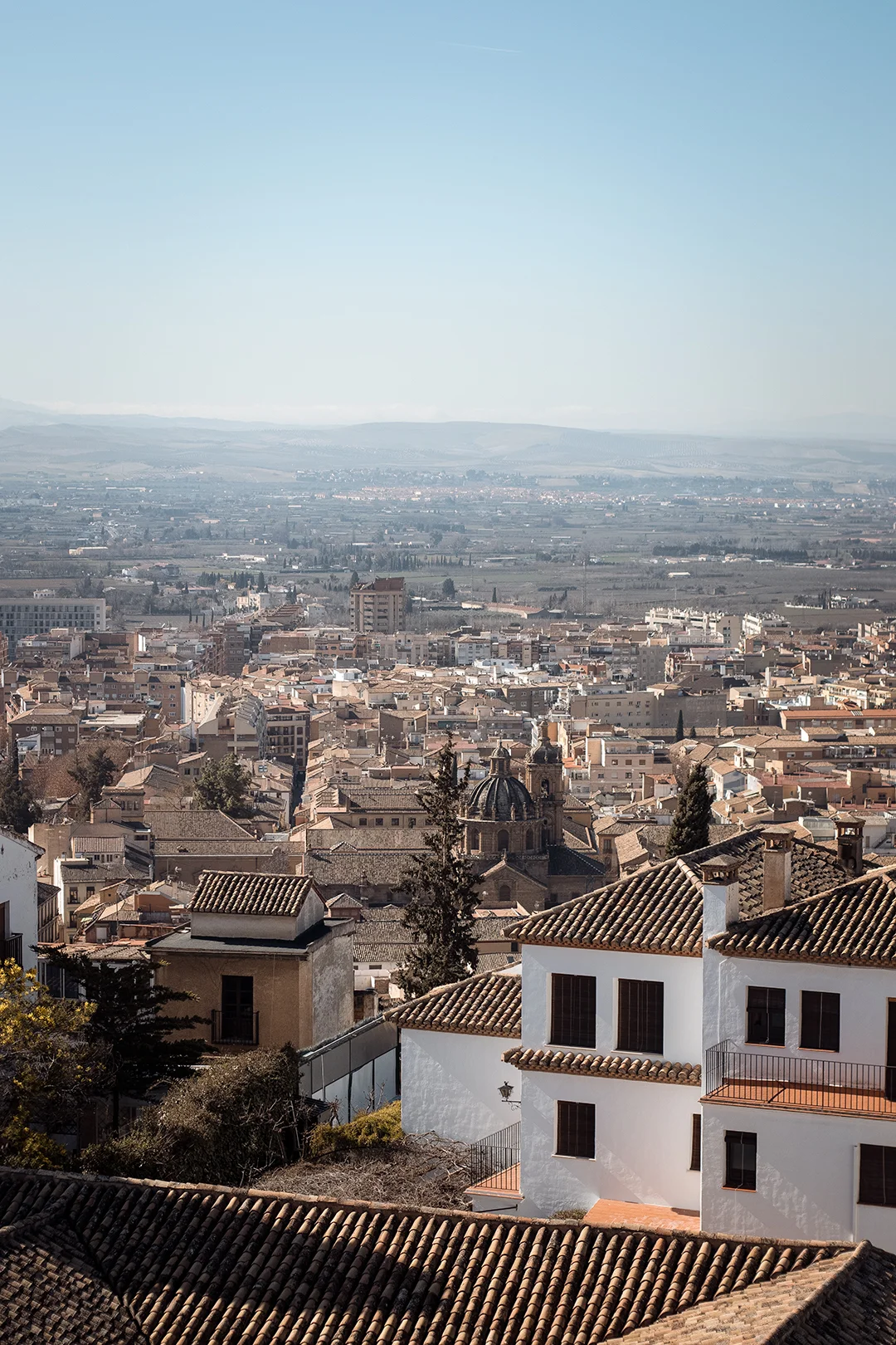 A sweeping panoramic view of Granada from the heights of the Albaicín — terracotta rooftops, the city cathedral dome, and the vast Vega de Granada stretching to the horizon.