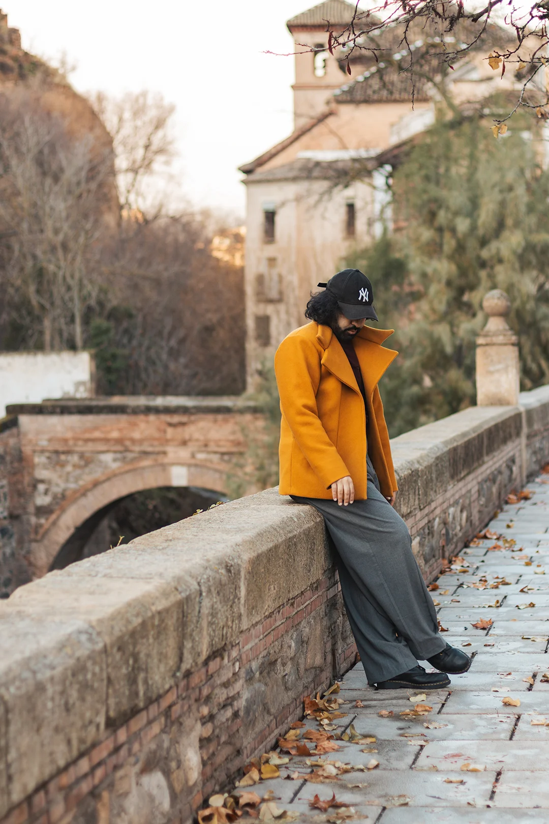 An individual portrait full of autumn mood — a young person leans on a centuries-old stone bridge in Granada's Río Darro area, golden leaves scattered on the cobblestones below.
