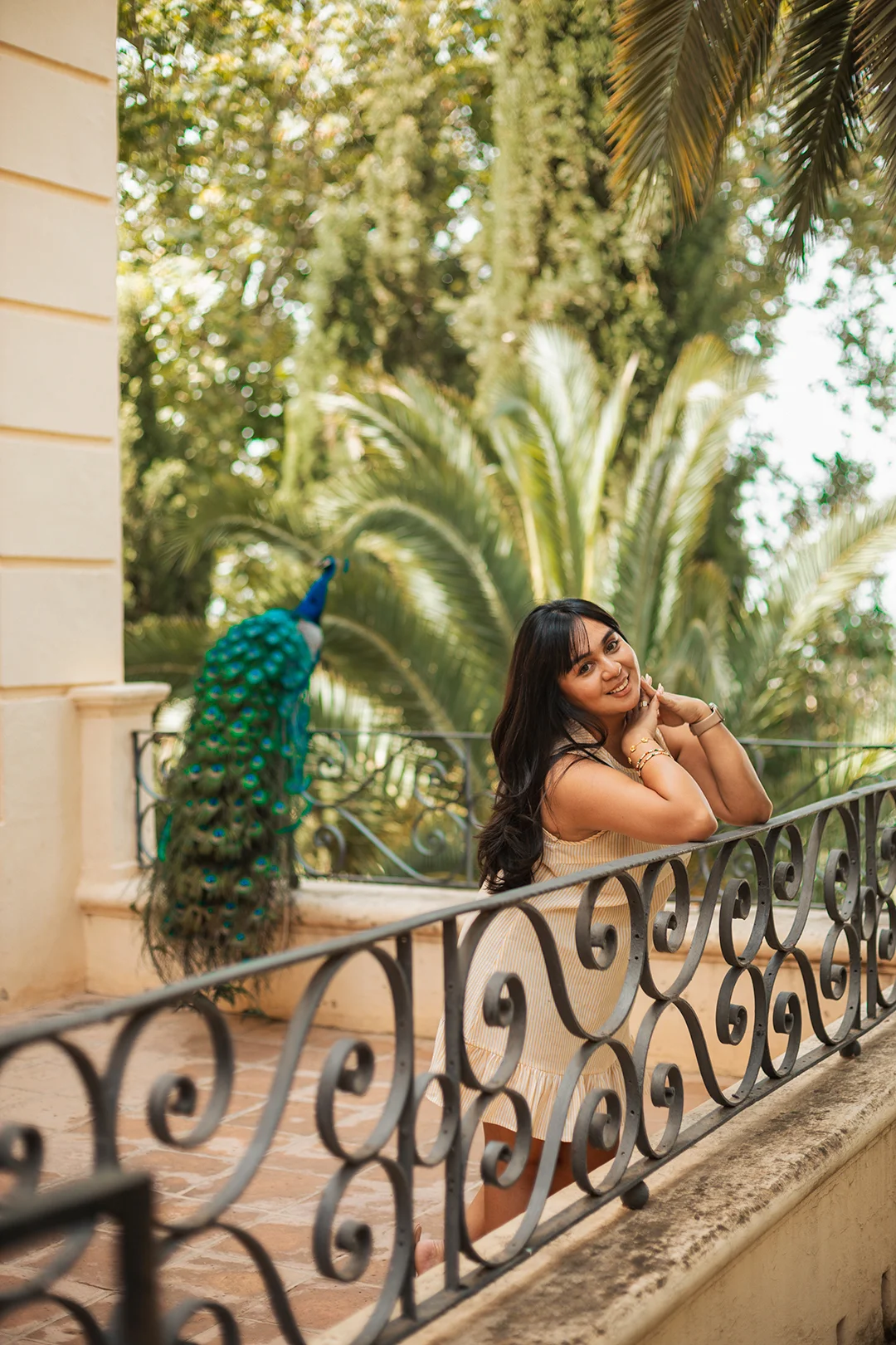 An enchanting individual portrait — a woman poses gracefully on a wrought-iron terrace railing with a magnificent peacock standing nearby, surrounded by a lush tropical garden in Andalusia.