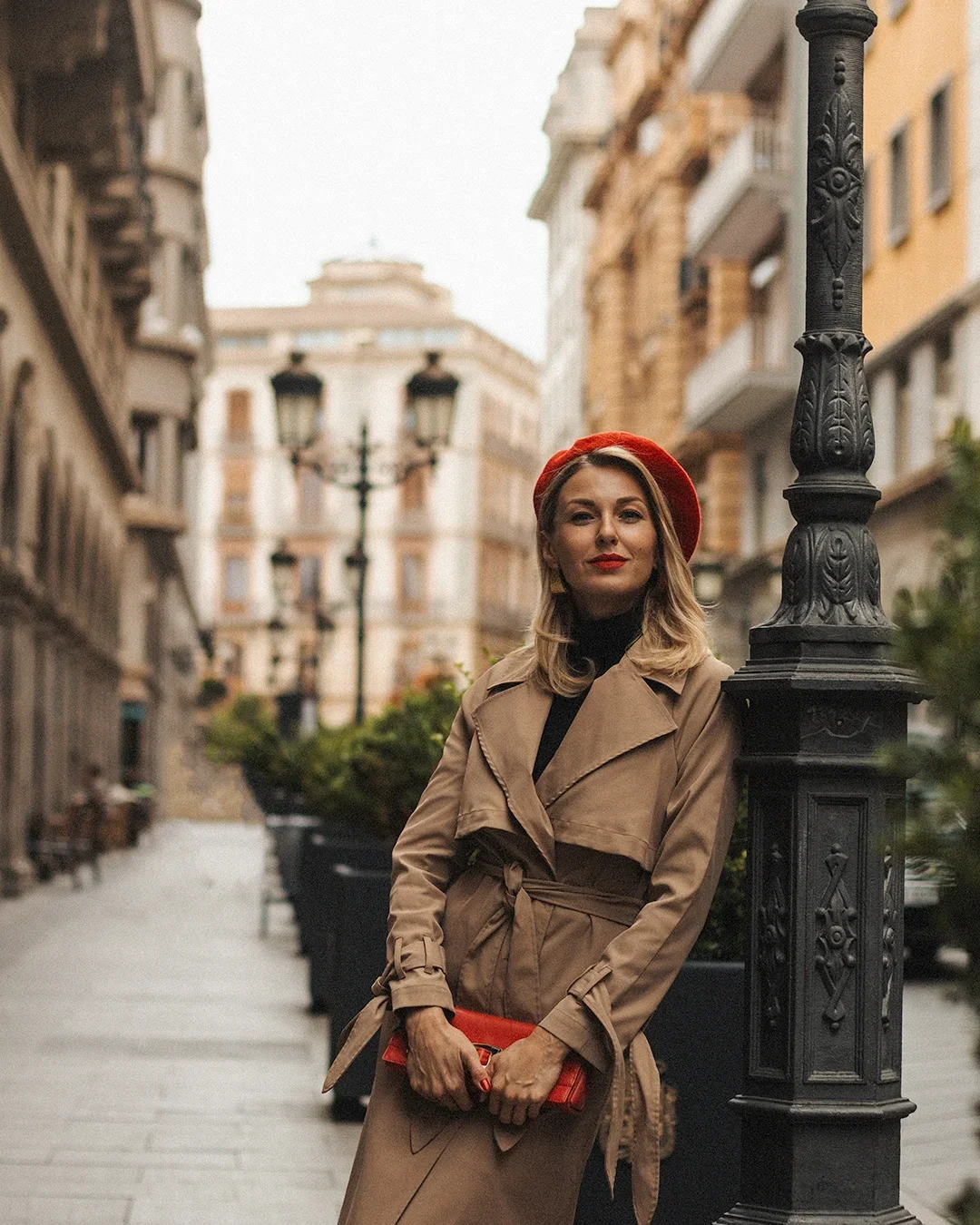 A chic fashion portrait — a blonde woman in a classic camel trench coat and bold red beret leans against a vintage street lamp on a quiet Andalusian city street, exuding effortless European style.