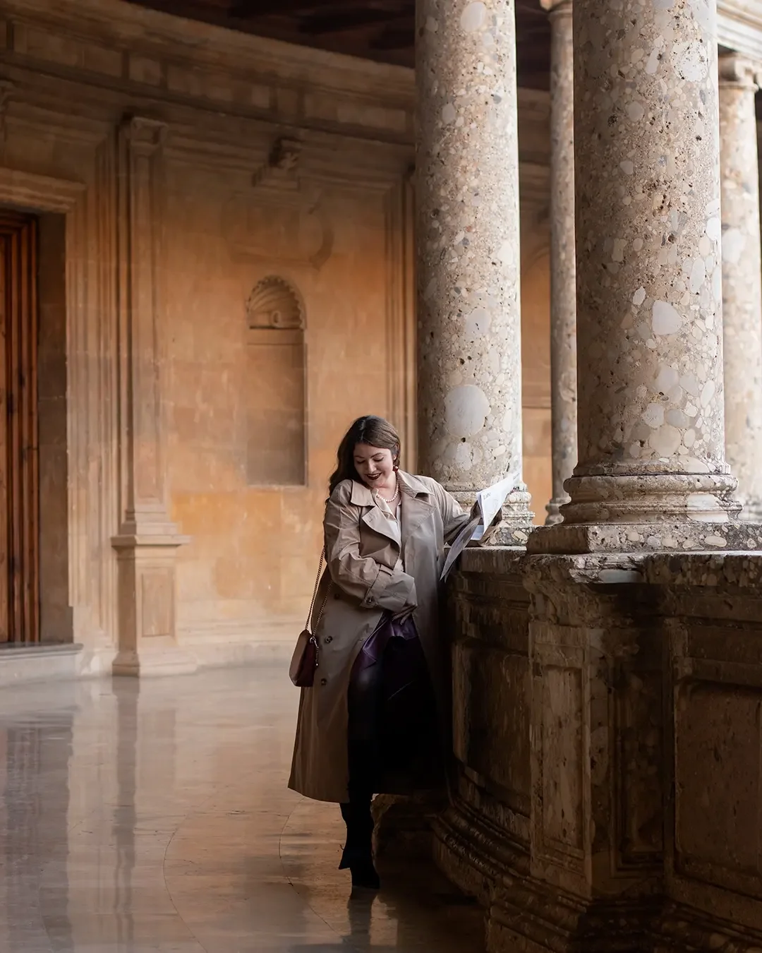 A poetic individual portrait — a woman in a camel trench coat reads quietly among the magnificent marble columns of the Palace of Charles V inside Granada's legendary Alhambra complex.