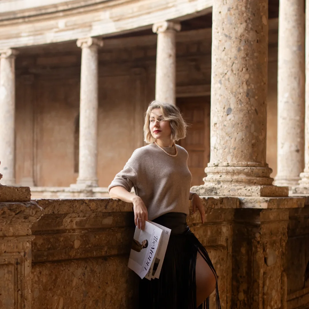 Timeless elegance — a sophisticated woman in a pearl necklace and grey cashmere sweater holds a fashion magazine among the ancient Roman columns of the Alhambra's Palace of Charles V, Granada.